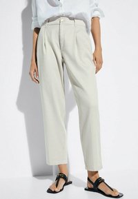 Pantaloni in lino beige chiaro con una vestibilità rilassata, dotati di pieghe e vita alta. Abbinati a sandali neri flat a piedi nudi.