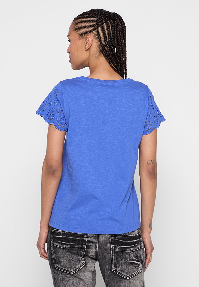 Blå t-shirt med korta ärmar och vågformig spetsdetalj på ärmarna, med en avslappnad passform och mjukt material. Bakifrån utan synliga varumärkesloggor.