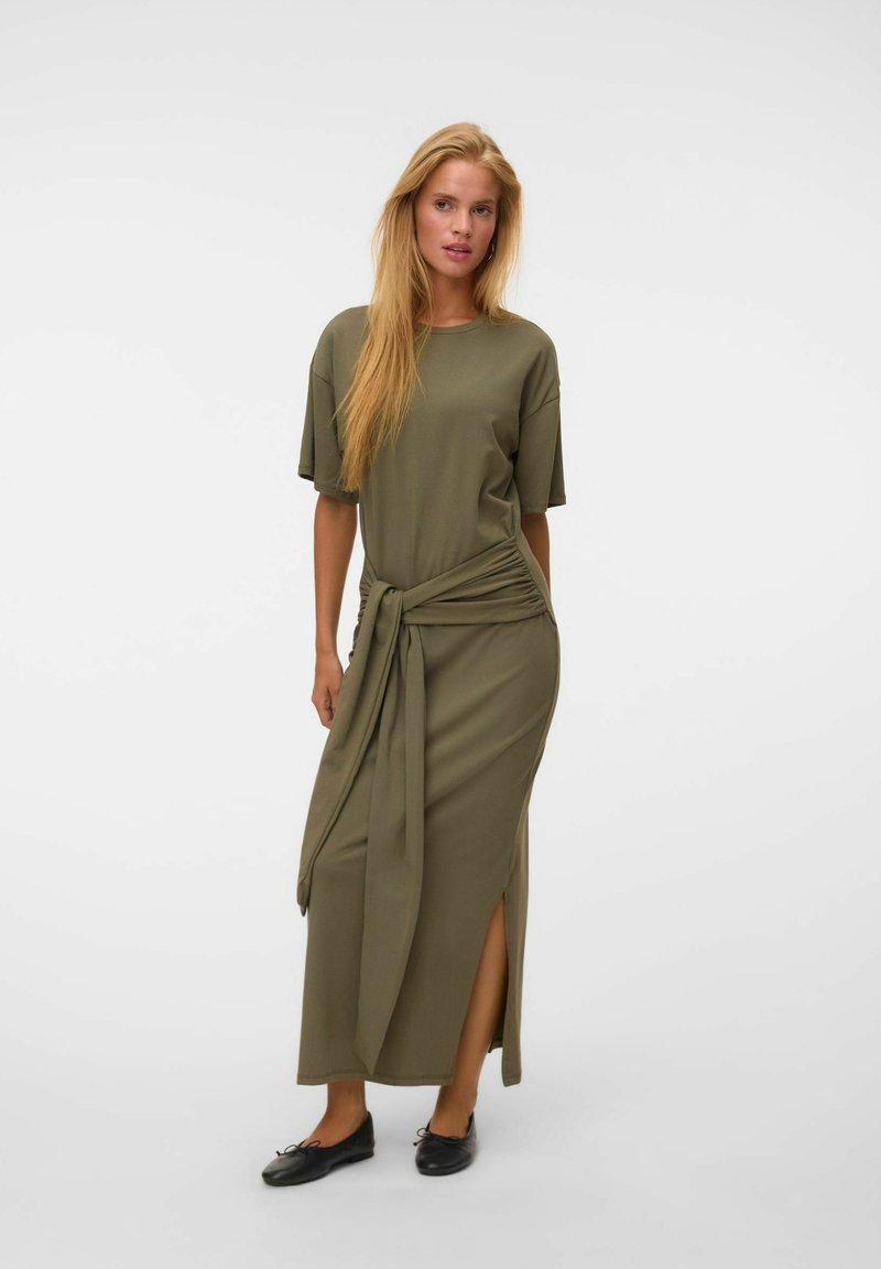 Femme blonde portant une robe midi vert olive à manches courtes, avec une fente latérale et un détail noué à l'avant, associée à des ballerines noires.