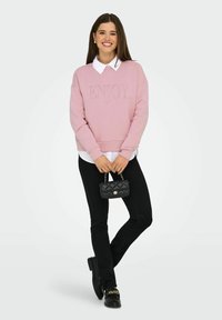 Glunderende vrouw staat in een roze trui met "ENJOY", een witte kraagshirt, zwarte broek, zwarte loafers met gouden kettingen en houdt een kleine zwarte handtas vast.