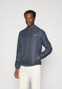 Sivá, ľahká bunda s kapucňou, zipsom, elastickými manžetami a zeleným logom Lacoste na prse; hladká, štruktúrovaná tkanina.