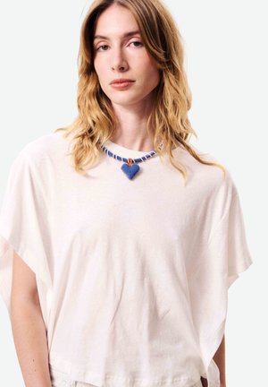 Jeune femme aux cheveux châtain clair ondulés, vêtue d'un t-shirt blanc ample et simple, portant un collier bleu perlé avec un pendentif en forme de cœur.