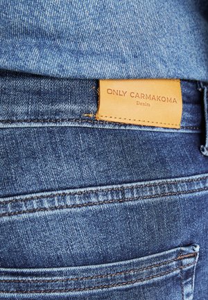Blå denimjeans med en brun läderetikett som läser "ONLY CARMKOMA Denim," med synlig sömnad och en sidofickdetalj.