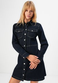 En blond kvinne iført en mørk denim skjortekjole med brystlommer, står mot en enkel hvit bakgrunn.