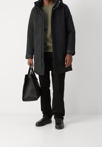 Svart fodrad parka med huva, med dragkedjestängning och gråa muddar. Svart texturerad axelväska. Grön sweatshirt och mörka byxor.