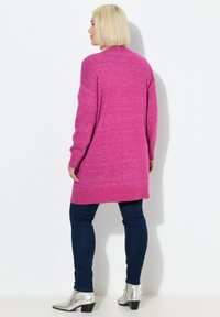 Cardigan lungo rosa con maglia strutturata, taglio comodo, polsini a costine e orlo uniforme, abbinato a jeans blu scuro e stivaletti argento.