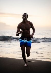 Zwarte zwembroek met blauwe en witte horizontale strepen, blote voeten, rennend op een zandstrand nabij de oceaan bij zonsondergang.