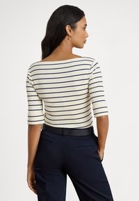 Žena s tmavými vlasmi, oblečená v bielej košeli s námorníckymi vodorovnými pruhmi a námorníckych nohaviciach, stojaca chrbtom, s jednou rukou v vrecku.