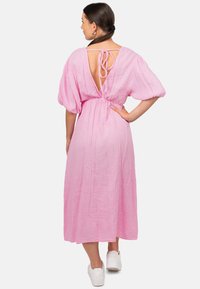 Roze, jurk van middellange lengte gemaakt van textuurstof; heeft een V-rug met strikdetail en pofmouwen. Gecombineerd met witte schoeisel.