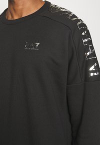 Felpa nera con scollo rotondo, caratterizzata da accenti riflettenti lucidi sulle spalle e un piccolo logo sul petto.