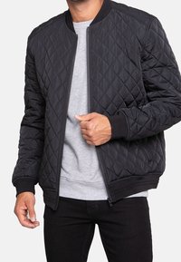Veste bombers matelassée noire avec poignets et col côtelés, dotée d'une fermeture éclair à l'avant et d'une coupe décontractée, portée par-dessus un sweat-shirt gris.