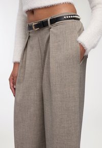 Pantaloni beige plissettati realizzati in un tessuto testurizzato, abbinati a una cintura nera con fibbia dorata e dettagli in perle lungo la vita.