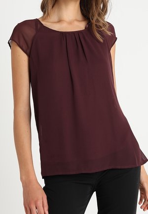 Femme portant un chemisier bordeaux foncé à manches courtes avec des plis au niveau de l'encolure, associé à un pantalon noir sur un fond blanc.