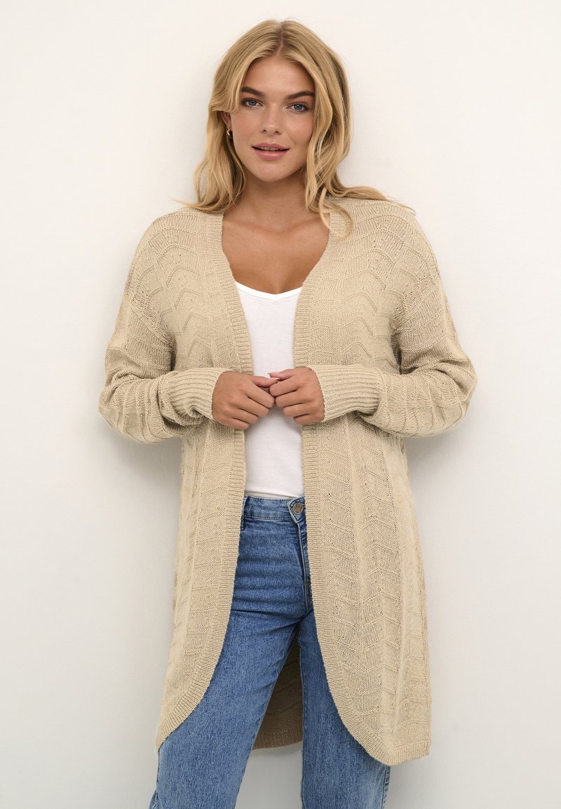 Beige pletený cardigan s chevron vzorem, dlouhými rukávy, otevřenou přední částí a volným střihem, kombinovaný s bílým topem a modrými džíny.