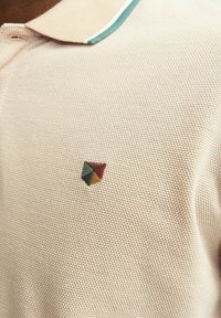 Polo tričko v svetlej béžovej farbe, s textúrovanou tkaninou. Obsahuje zelený lem goliera a na hrudi má viacfarebný trojuholníkový logo.