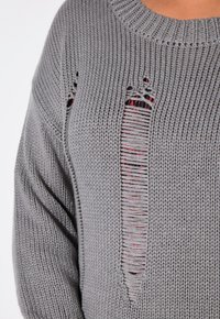 Grauer Strickpullover mit weitem Ausschnitt, der markante vertikale Schlitze aufweist, die das darunterliegende rot-schwarze Muster enthüllen.