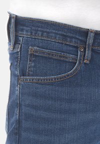 Jeans i denim med en mörkblå färg, prydda med kopparfärgad sömnad, en frampocket och dragkedja. Materialet verkar robust och texturerat.