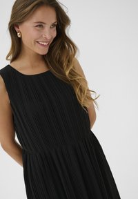 Svart plisserad klänning med rund halsringning och ärmlös design, med en midjesöm. Materialet verkar slätt med vertikal textur.
