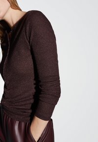 Donna che indossa un top lungo a maniche con brillanti di colore marrone scuro e pantaloni in pelle nera, con le mani in tasca, su uno sfondo chiaro e uniforme.