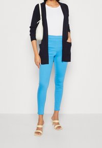 Granatowy kardigan z kieszeniami, biały T-shirt, jasnoniebieskie dopasowane dżinsy oraz beżowe sandały na obcasie. Gładkie faktury i kontrastujące kolory.