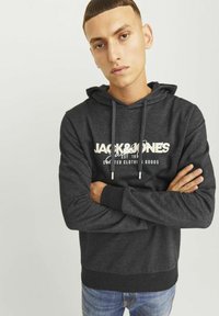 Donkergrijze hoodie met een trekkoord, voorzien van een wit logo op de voorkant, geribbelde manchetten en een casual pasvorm. Draagt met spijkerbroek.