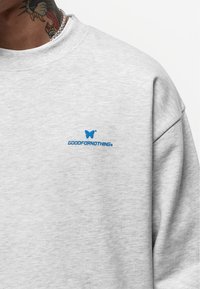 Lichtgrijze sweater met ronde hals, met een blauw vlinderlogo en de tekst "GOODFORNOTHING" geborduurd op de borst. Zachte textuur.