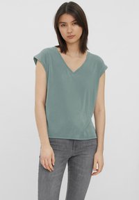 Kurzärmliges V-Ausschnitt-T-Shirt aus weichem, sanften grünem Stoff. Lässige Passform mit umgeschlagenen Ärmeln und glatter Textur. Kombiniert mit grauen Jeans.