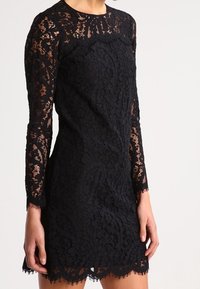 Kobieta ubrana w dopasowaną czarną koronkową sukienkę z długimi przezroczystymi rękawami i falbaniastym brzegiem, stojąca na jednolitym jasnym tle.
