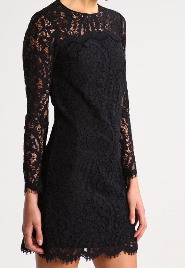 Kobieta ubrana w dopasowaną czarną koronkową sukienkę z długimi przezroczystymi rękawami i falbaniastym brzegiem, stojąca na jednolitym jasnym tle.