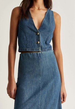 Vrouw die een mouwloos, kort denimvest met twee knopen en een hoge taille denimrok draagt, neutrale achtergrond.