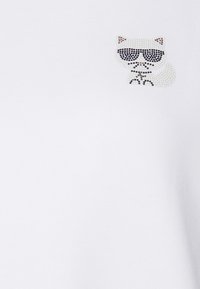 La t-shirt bianca presenta un design di un gatto con occhiali da sole in strass sul lato sinistro del petto. Il materiale appare liscio e simile al cotone nella consistenza.