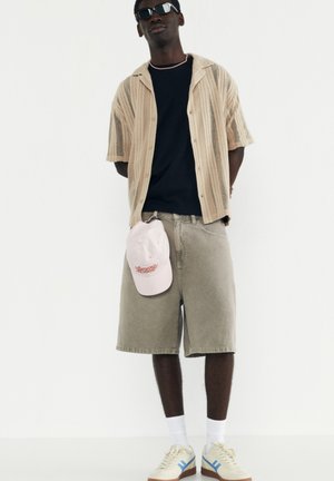 Beige knit short-sleeve shirt, black t-shirt, gray shorts, pink cap, and beige sneakers with blue accents; dark sunglasses; standing pose.