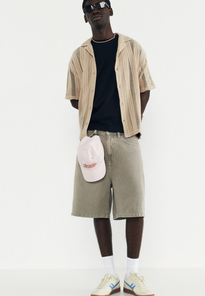 Beige strik med korte ærmer, sort t-shirt, grå shorts, lyserød kasket og beige sneakers med blå detaljer; mørke solbriller; stående pose.