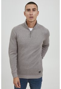 Pull en tricot gris clair avec un col zippé au quart, des poignets et un ourlet côtelés. Présente un motif texturé et une étiquette de logo en bas à gauche.