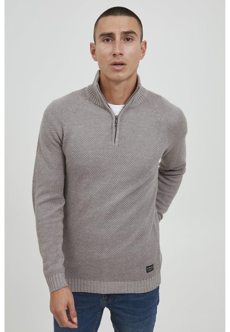 Pull en tricot gris clair avec un col zippé au quart, des poignets et un ourlet côtelés. Présente un motif texturé et une étiquette de logo en bas à gauche.