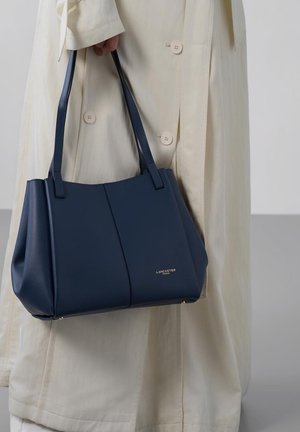 Persoon in beige jas die een navy blauwe leren Lancaster Paris handtas vasthoudt aan twee schouderbanden.