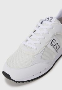 Witte sportieve schoen van mesh en synthetisch materiaal, met een zwarte buitenzool, witte veters en een zwart "EA" logo aan de zijkant.