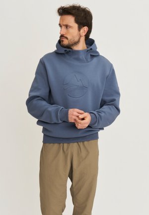 Homme barbu portant un sweat à capuche bleu et un pantalon beige, regardant sur le côté avec les mains jointes devant, debout devant un fond neutre.