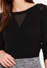 Svart långärmad topp med ett genomskinligt triangulärt panel vid halsringningen, texturerad tyg och en något figurnära design.