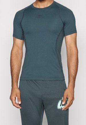 Mężczyzna noszący dopasowaną ciemnoturkusową koszulkę Nike Pro z krótkim rękawem oraz dopasowane sportowe spodnie z logo Nike na lewym udzie.
