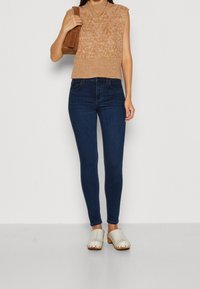 Světle hnědý bezrukávový pletený svetr s texturovaným vzorem, tmavě modré skinny džíny a béžové dřeváky s dřevěnými podrážkami. Hnědá kabelka na rameno.