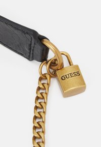 Sort tekstureret rem med en gylden metal kæde og hængelås. Hængelåsen har et prægede "GUESS"-logo, der tilføjer et brandet accent.