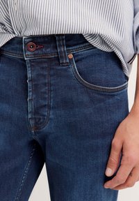 Dunkelblaue Jeans mit hoher Taille, fünf Taschen, minimalen Nähdetails und einem einzelnen Knopfverschluss.