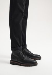 Czarne skórzane botki za kostkę z białym przeszyciem, okrągłym noskiem, sznurowanym zapięciem i wytrzymałą podeszwą, zestawione z podwiniętymi czarnymi spodniami.