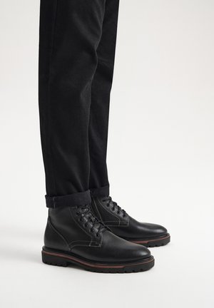 Czarne skórzane botki za kostkę z białym przeszyciem, okrągłym noskiem, sznurowanym zapięciem i wytrzymałą podeszwą, zestawione z podwiniętymi czarnymi spodniami.