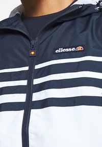 Veste zippée marine et blanche avec des rayures horizontales, un col montant et un petit logo de marque brodé sur la poitrine.