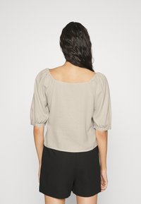 Ljusbeige puffärmad topp med fyrkantig halsringning, tillverkad av ett texturerat tyg. Baksidan är enkel med en kortad längd. Matchas med svarta shorts.