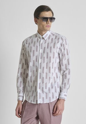 Man draagt een wit overhemd met bruine verticale strepen, mauve broek en grote donkere zonnebril, kijkt opzij.