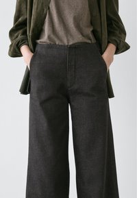 Pantalon à jambes larges en tissu foncé avec une texture subtile. Il comprend des poches latérales et une taille ajustée. Porté avec un haut gris et une veste vert foncé.