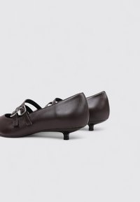 Zapatos de cuero marrón con puntera redonda, tacón pequeño y detalles de correas con hebillas metálicas. Textura suave con acabado brillante.